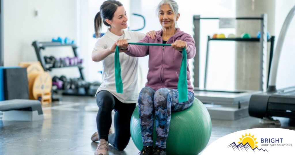 A care provider helps an older woman exercise with a resistance band and an exercise ball, representing how exercise can help with senior fall prevention.