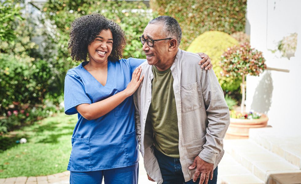Companion caregiver smiling and walking beside a senior man outdoors, reflecting compassionate Companionship Care in Whitewater that promotes connection and independence.